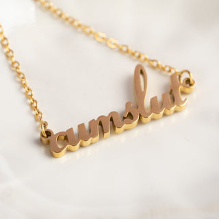 A gold cumslut necklace elegantly draped on a surface, with the pendant resting prominently. The delicate chain flows naturally, highlighting its shine and intricate design.