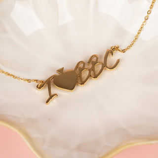 A gold "I Heart BBC" necklace with bold lettering and a heart symbol is elegantly draped across a glossy white display tray.