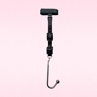 A black adjustable restraint harness with a padded top pad, multiple buckle straps, and a metal J-hook hanging from the bottom, shown against a light pink textured background.