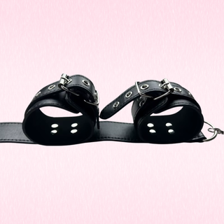 A pair of black faux-leather wrist cuffs with silver metal grommets and buckles, connected by a black strap, shown against a solid light pink background.