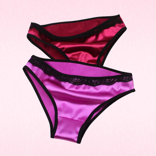 Two satin briefs with black lace trim and black edging, one in red and one in purple, displayed against a light pink background.