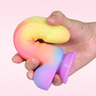 A hand holding a flexible, U-shaped silicone dildo with a pastel rainbow gradient of yellow, pink, orange, blue, and purple, against a solid light pink background.