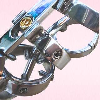 A close-up of a polished chrome metal screws, rings, and a small gold-colored adjustment screw, set against a solid light pink background.