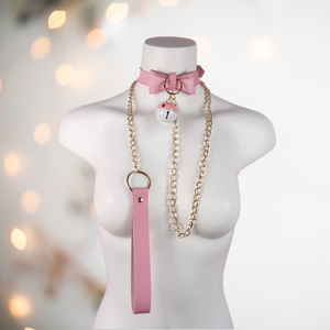 A pink choker neck collar with buckle fastening, has a decorative bow to the front with large pink and white bell, it also comes with a gold chain lead. 