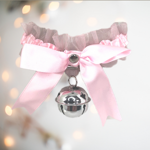 A satin and gauze fabric choker / necklace that has a matching satin bow to the front, with a large hanging bell. There are ribbon string ties to the back, allowing for different sized necks and it comes in soft pink.