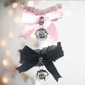 A satin and gauze  fabric choker / necklace that has a matching satin bow to the front, with a large hanging bell. There are ribbon string ties to the back, allowing for different sized necks and it comes in soft pink or black. 