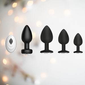 The image shows the vibrating anal plug with its remote control and the three other non vibrating anal plugs in small, medium and large. They are black with a diamond style crystal base gem stone.